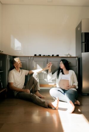 Pregnant couple sitting on kitchen floor, touching hands, in a sunlit home interior. Peaceful and intimate setting.